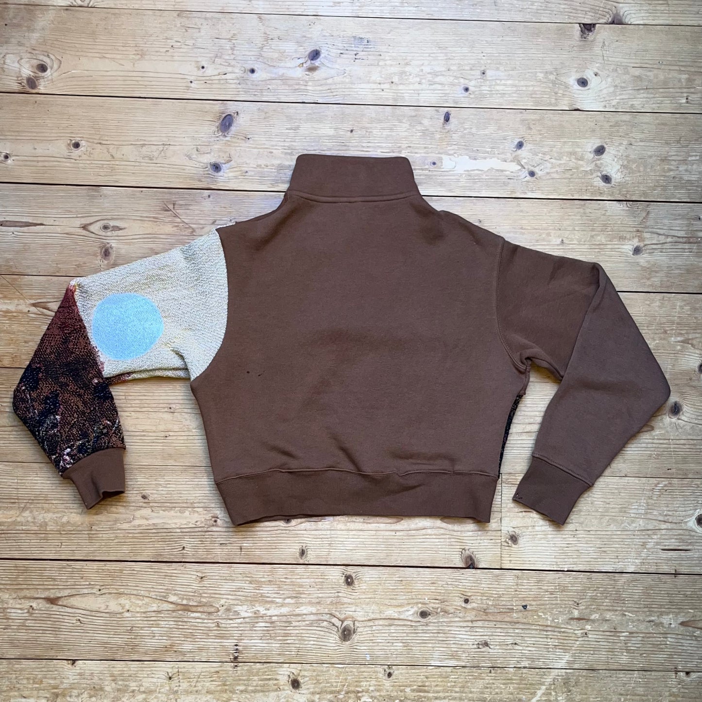 Rückansicht des braunen Recrafted Half-Zip Pullovers von Sun Seam Studio mit Solar Flower Stoffpanel am Ärmel, flach liegend auf Holzboden