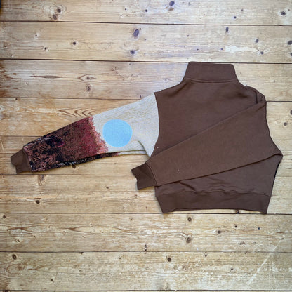 Seitenansicht des braunen Recrafted Pullovers mit ausgestrecktem Ärmel – Solar Flower Motiv von Sun Seam Studio in voller Länge sichtbar, Einzelstück auf Holzboden