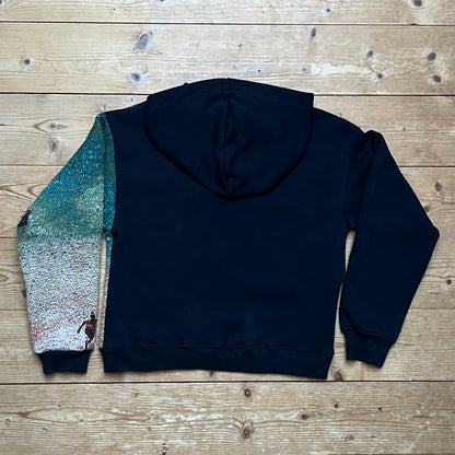 Rückansicht des Recrafted Beach Days Hoodie – schwarzer Cropped Hoodie mit Kapuze, rechter Ärmel mit handvernähtem Strandmotiv im Farbverlauf, flach liegend auf Holzuntergrund